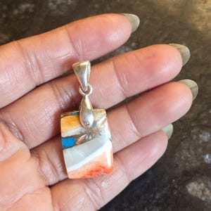 May include: Rectangular pendant with a silver bail. The pendant features a colorful stone with orange, white, blue, and gold hues. The stone is set in a silver bezel. The pendant is held in a hand.