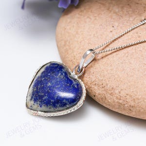 May include: A heart-shaped pendant with a deep blue gemstone, set in a silver-colored metal frame. The pendant hangs from a delicate silver chain. The gemstone has a speckled appearance. The jewelry is displayed on a light brown stone.