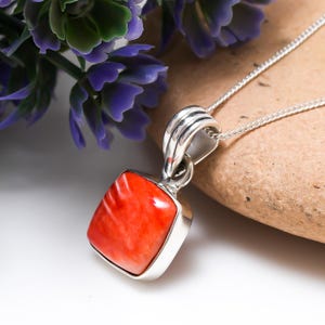 Könnte beinhalten: Quadratischer Anhänger aus Silber mit einem leuchtend orangefarbenen Stein. Der Stein ist in eine silberne Fassung gefasst und hängt an einer feinen Silberkette. Der Stein hat ein einzigartiges, wirbelndes Muster. Im Hintergrund sind lila Blumen und eine hellbraune Oberfläche.