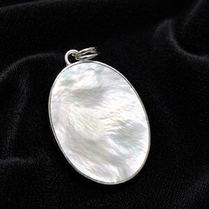May include: Oval pendant with a pearlescent, white surface and a silver-colored metal frame. The pendant has a loop at the top for a chain. The background is a black, textured fabric.