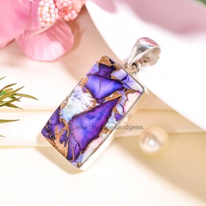 Op de afbeelding: Een rechthoekige hanger met een levendige paarse en gouden steen, gezet in een zilveren frame. De steen heeft een uniek patroon met tinten paars, wit en goud. De hanger heeft een zilveren beugel om aan een ketting te bevestigen. De tekst "Jewelry onesandgems" is zichtbaar.