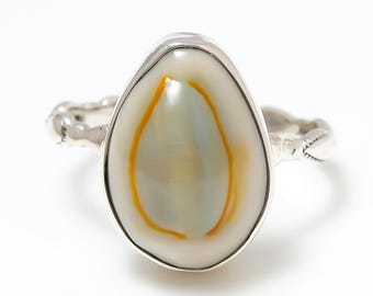 Könnte beinhalten: Silberring mit einem großen, ovalen Muschelmittelstück. Die Muschel hat eine cremeweiße Basis mit einem gelb-orangen Rand und einem blassblau-grünen Zentrum. Das Band hat ein strukturiertes Design.