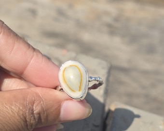 Könnte beinhalten: Silberring mit einem großen, ovalen, cremefarbenen Stein mit einem dünnen, gelb-orangen Streifen. Das Band des Rings hat ein strukturiertes Design. Der Stein ist in eine silberne Lünette gefasst.