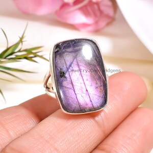 May include: A silver ring featuring a large, rectangular, purple labradorite gemstone. The stone has a shimmering, iridescent quality with streaks of lavender and violet. The ring's band is a simple, split-shank design.