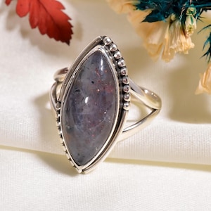 May include: A silver ring featuring a large, oval-shaped gemstone with a mix of purple, gray, and red hues. The gemstone is set in a detailed silver band with a beaded edge. The ring's design is elegant and eye-catching.