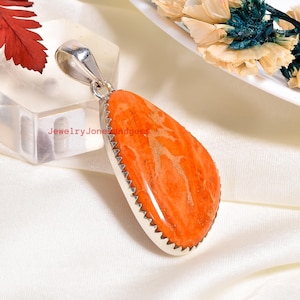 May include: Orange gemstone pendant with a silver bail and a serrated silver edge. The pendant has a teardrop shape and a natural, textured appearance. The text "JewelryJonesandgems" is visible on the pendant.