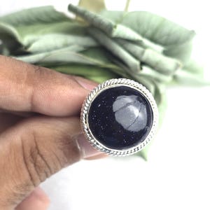 May include: A silver ring featuring a large, round, dark blue stone with sparkling inclusions. The stone is set in a silver bezel with a twisted rope design. The ring is set against a backdrop of green leaves.