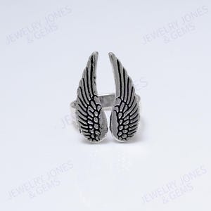 Puede incluir: Un anillo plateado con dise&ntilde;o de alas. El anillo tiene dos alas con patrones de plumas detallados. El anillo est&aacute; sobre un fondo blanco. El texto "JEWELRY JONES & GEMS" es visible en la imagen.