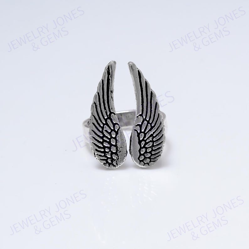 Puede incluir: Un anillo plateado con dise&ntilde;o de alas. El anillo tiene dos alas con patrones de plumas detallados. El anillo est&aacute; sobre un fondo blanco. El texto "JEWELRY JONES & GEMS" es visible en la imagen.