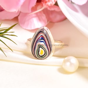 May include: A silver ring with a large, teardrop-shaped stone. The stone has a unique pattern of layered colours including black, red, blue, and yellow. The ring is set against a soft, white background with a pearl and pink flowers.