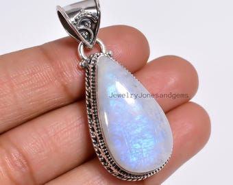 Pode incluir: Um pingente de pedra da lua em forma de gota com uma argola prateada. A pedra da lua tem um brilho perolado e iridescente. O pingente é segurado por uma mão. O texto "JewelryJonesandgems" é visível.