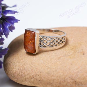 May include: A silver ring with a rectangular, orange-brown gemstone. The band features an open, woven design. The ring is displayed on a light brown, textured surface. The words "JEWELRY JONES & GEMS" are visible in the background.
