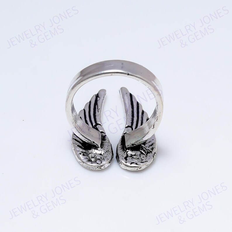 Puede incluir: Un anillo plateado con dise&ntilde;o de alas. El anillo presenta dos alas detalladas con patrones de rayas, sobre un fondo blanco brillante. La banda del anillo es lisa y redondeada.