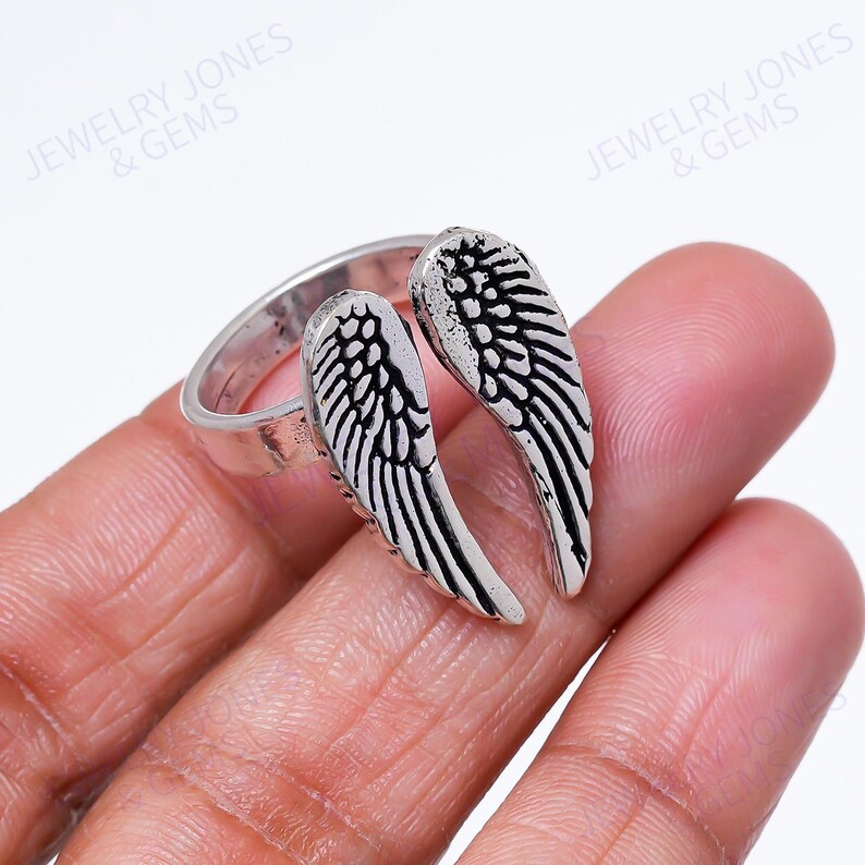 Puede incluir: Un anillo plateado con un dise&ntilde;o de alas detallado. El anillo tiene dos alas que se curvan alrededor del dedo. Las alas est&aacute;n intrincadamente detalladas con patrones de plumas. El anillo se muestra sobre un fondo claro. Joyer&iacute;a.
