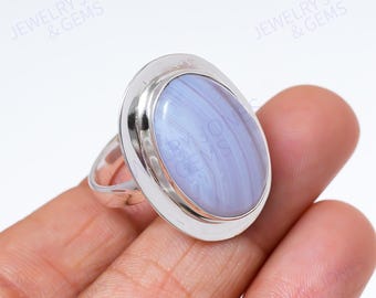 Könnte beinhalten: Ein Silberring mit einem großen, ovalen Edelstein aus blauem Spitzenachat. Der Stein zeigt ein weiches, gebändertes Muster in Hellblau und Weiß. Der Ring hat ein schlichtes, poliertes Silberband und eine Lünettenfassung.