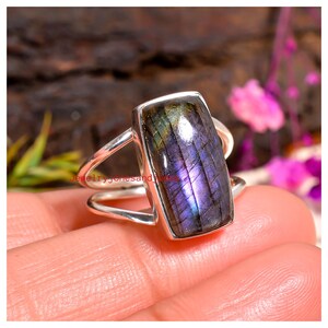 Puede incluir: Anillo de plata con una piedra labradorita rectangular. La piedra muestra tonos de púrpura, azul y verde. El anillo tiene un diseño de caña dividida. El texto "jewelryjonesandmore" es visible.