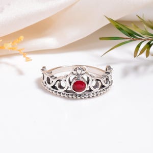 Op de afbeelding: Een zilverkleurige kroonring met een centrale rode edelsteen. De ring heeft een gedetailleerd ontwerp met een reeks kleine, decoratieve kralen langs de basis en ingewikkelde opengewerkte patronen. De ring staat tegen een witte achtergrond met zachte verlichting.
