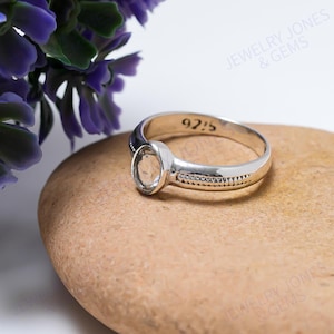 Könnte beinhalten: Ein silberner Ring mit einem klaren, ovalen Edelstein. Das Ringband hat ein strukturiertes Design mit kleinen, eingebetteten Steinen. Die Zahl "925" ist auf dem Band eingraviert. Der Ring wird auf einem hellbraunen Stein präsentiert.