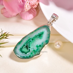 May include: A pendant featuring a polished, irregularly shaped green agate stone with a silver-colored bail. The agate has a vibrant green center with white and light green banding. The pendant is displayed on a white surface with a pink flower and a pearl.