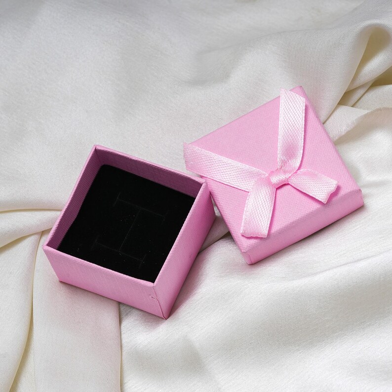 Puede incluir: Caja de joyer&iacute;a rosa abierta, con interior de terciopelo negro. La tapa est&aacute; atada con una cinta rosa. La caja est&aacute; sobre una tela blanca y sedosa.