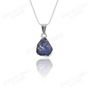 Op de afbeelding: Een zilveren hangerketting met een ruwe, blauw-paarse edelsteen. De hanger heeft een onregelmatige vorm en is gezet in een zilveren bezel. De ketting hangt aan een fijne zilveren ketting. De achtergrond is wit.