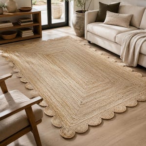 May include: A rectangular area rug crafted from natural tan jute fibers, featuring a braided texture and a decorative scalloped edge border. The rug is woven in a concentric rectangular pattern, showcasing the organic light brown color of the material.