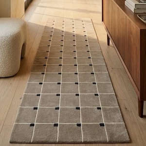 May include: A long rectangular runner rug featuring a grid pattern of light brown squares separated by thin white lines and small black squares at each intersection. The rug is positioned on a light wood floor in a hallway with a cream-colored ottoman.