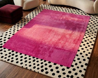 May include: A rectangular area rug featuring a central block of vibrant pink and magenta ombre coloring, surrounded by a wide border of cream-colored fabric decorated with a repeating pattern of irregular black polka dots across the surface.