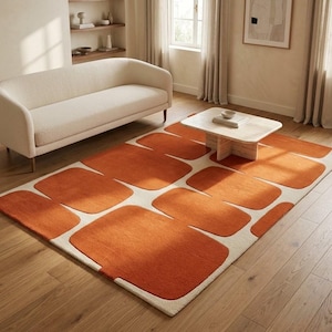 May include: A rectangular area rug featuring a repeating pattern of rounded, burnt orange rectangular shapes set against a cream-coloured background. The rug has a textured, pile surface and covers a portion of the wooden floor in the room.
