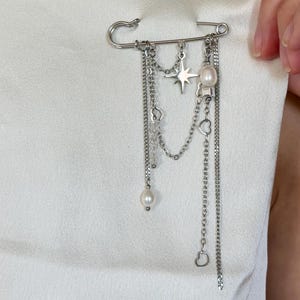 May include: A silver safety pin brooch with dangling chains, a star, pearls, and heart charms. The brooch is attached to a white garment, showcasing its decorative design and intricate details. This accessory adds a touch of elegance.