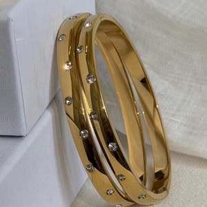 May include: Two polished gold-tone hinged bangle bracelets featuring a series of small, clear, round-cut embedded crystals spaced evenly along the outer surface of each band. The bracelets have a smooth, reflective finish and a secure clasp mechanism.