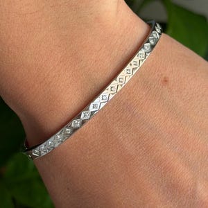 May include: A silver-toned metal bangle bracelet featuring a repeating geometric pattern of diamond-shaped cutouts. Each diamond shape contains a small, clear, sparkling stone set within the centre of the metallic frame, creating a continuous band.