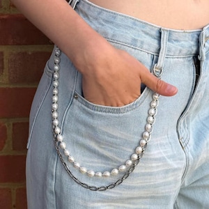 Puede incluir: Una cadena de cartera de doble capa que presenta una hebra de perlas blancas, redondas y brillantes, y una hebra de eslabones ovalados de metal plateado. La cadena está sujeta a la trabilla de un pantalón vaquero azul claro con un cierre circular.