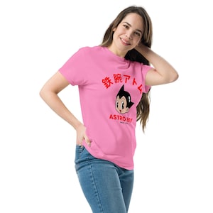 May include: Pink t-shirt featuring a graphic of Astro Boy with Japanese text and the words "ASTRO BOY SINCE 1952" in red. The t-shirt is made of a soft material.