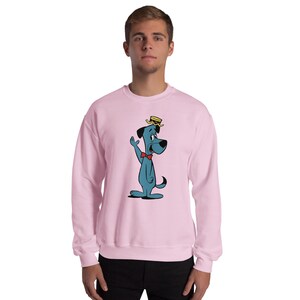 Op de afbeelding: Een lichtroze sweatshirt met een cartoonhond in blauw, met een rode vlinderdas en een gele hoed. De hond zwaait. De sweatshirt heeft een ronde hals.