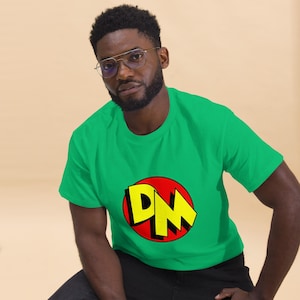 May include: A green t-shirt with a circular graphic. The graphic features the letters "DM" in yellow against a red background. The t-shirt is made of a soft material, suitable for casual wear.