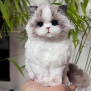 May include: A plush, fluffy cat toy with a white body, gray and white markings on its head, and large, dark eyes. The toy cat has a pink nose and a gray tail. The cat is sitting upright, and the background is blurred green foliage.