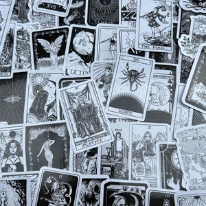 May include: A collection of black and white tarot cards featuring various illustrations and text. The cards depict celestial bodies, figures, and symbols, including "The Star," "The Fool," and "Justice." The cards are scattered across a surface.