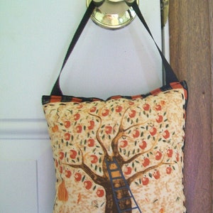 May include: A decorative pillow with a black strap hanging from a door knob. The pillow features a tree with red apples, a blue ladder, and a pumpkin. The pillow has a beige background with a black and orange border.