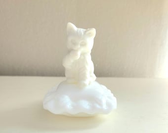 May include: White milk glass figurine of a cat sitting on a cloud. The cat is holding its paw to its mouth.