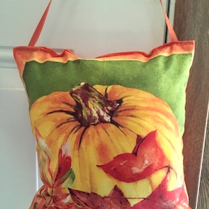 May include: A decorative pillow featuring a watercolor painting of a large pumpkin with a green background and autumn leaves. The pillow has an orange ribbon for hanging. The color palette includes shades of orange, yellow, red, and green.