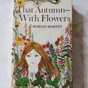 May include: A hardcover book titled That Autumn—With Flowers by Marian Martin, featuring an illustration of a person with long brown hair surrounded by colorful flowers, including yellow daisies, pink roses, and green ferns on a cream background.
