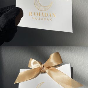 May include: White gift bag with a gold satin bow. The bag features a gold crescent moon design and the words "RAMADAN MOBARAK" in gold lettering. The bag is held by a person wearing a black glove.