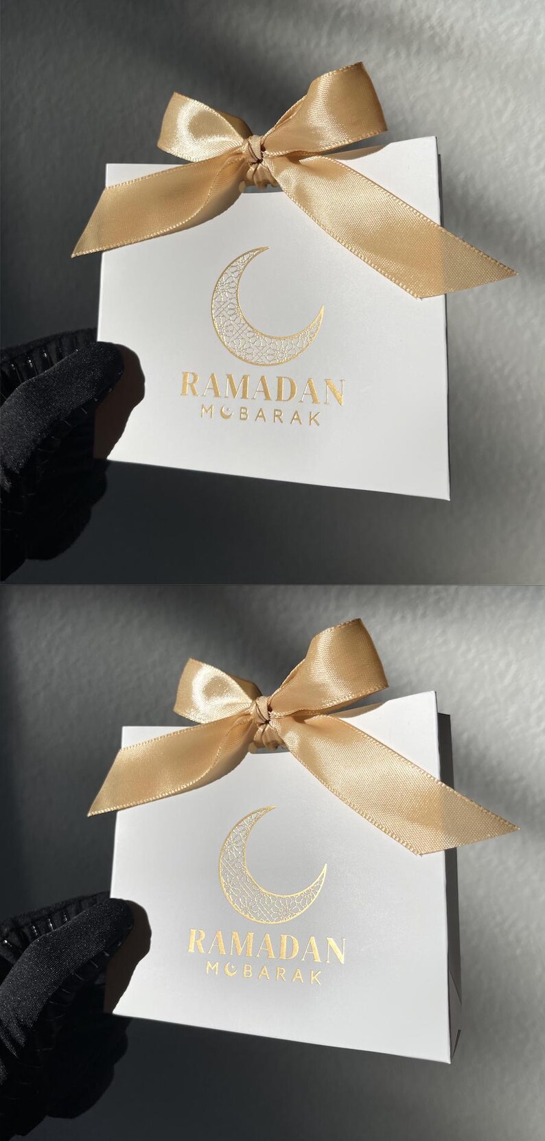 May include: White gift bag with a gold satin bow. The bag features a gold crescent moon design and the words "RAMADAN MOBARAK" in gold lettering. The bag is held by a person wearing a black glove.