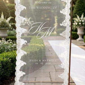 K&ouml;nnte beinhalten: Ein hohes, rechteckiges, transparentes Acryl-Hochzeitsschild mit goldfarbenem Metallrahmen und wei&szlig;er Spitzenborte an den R&auml;ndern. Der Text lautet: Welcome to THE WEDDING OF, das Monogramm HM, HEIDI & MAXWELL und das Datum 06. 02. 2028.