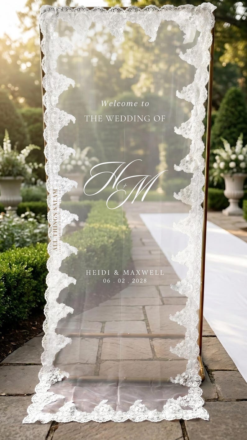 K&ouml;nnte beinhalten: Ein hohes, rechteckiges, transparentes Acryl-Hochzeitsschild mit goldfarbenem Metallrahmen und wei&szlig;er Spitzenborte an den R&auml;ndern. Der Text lautet: Welcome to THE WEDDING OF, das Monogramm HM, HEIDI & MAXWELL und das Datum 06. 02. 2028.