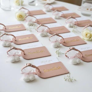 May include: A collection of pink luggage tags with the gold text "Bismillah" and small white favor bags tied with pink ribbons. White cards with "Thank You" are also present, along with white roses and baby's breath.