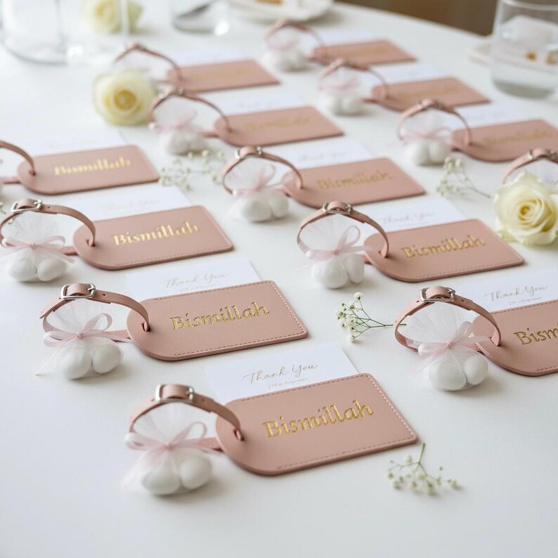 May include: A collection of pink luggage tags with the gold text "Bismillah" and small white favor bags tied with pink ribbons. White cards with "Thank You" are also present, along with white roses and baby's breath.