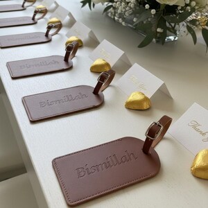 Pu&ograve; includere: Etichette per bagagli in pelle marrone con la scritta "Bismillah" in rilievo sono disposte su una superficie bianca. Sono presenti anche piccoli biglietti bianchi con testo dorato e cioccolatini incartati in oro. Un bouquet di rose bianche e velo da sposa &egrave; sullo sfondo.
