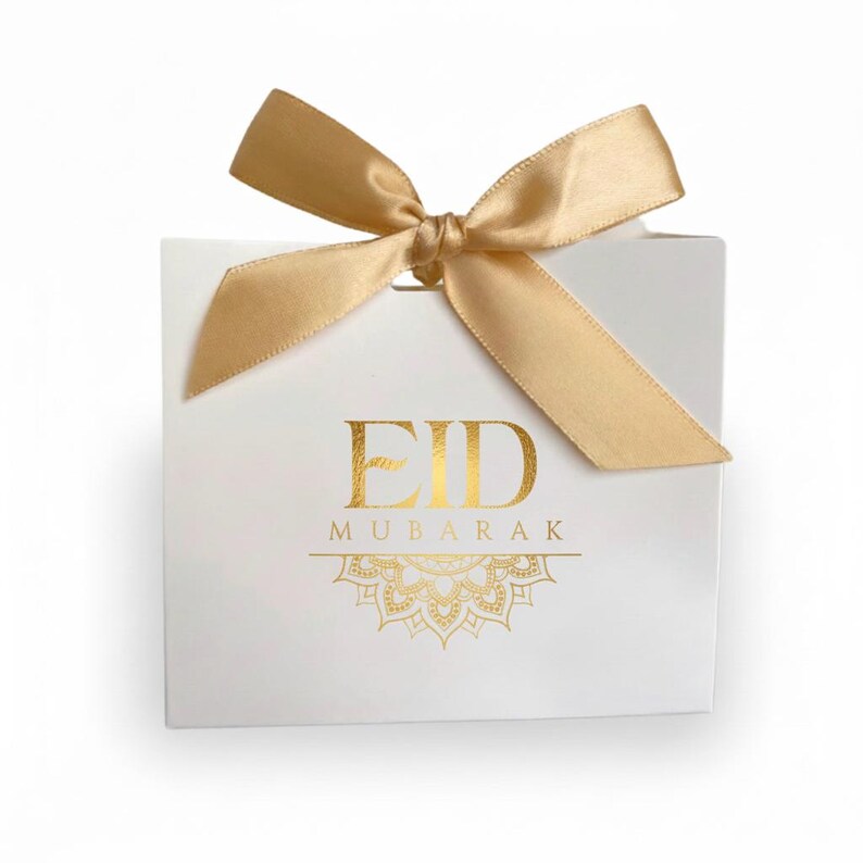 K&ouml;nnte beinhalten: Eine wei&szlig;e Geschenkbox mit goldfarbenem Schriftzug "EID MUBARAK" und einem dekorativen Golddesign. Die Box ist mit einer goldenen Satin-Schleife gebunden.
